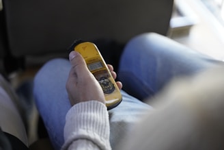 A person is holding a yellow handheld device, possibly a measuring instrument or meter, while sitting on a chair. The person is wearing a light-colored sweater and jeans. The image is slightly out of focus, creating a soft background.