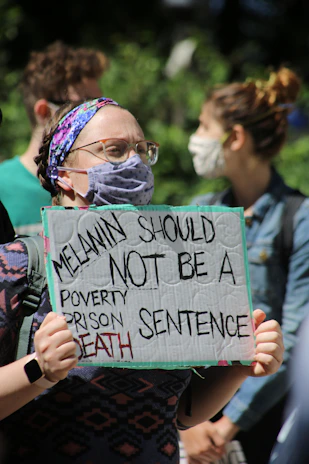 A person wearing a face mask and a colorful headband holds a protest sign that reads 'Melanin should not be a poverty, prison, death sentence'. Others in the background also wear masks, indicating a public gathering or protest.