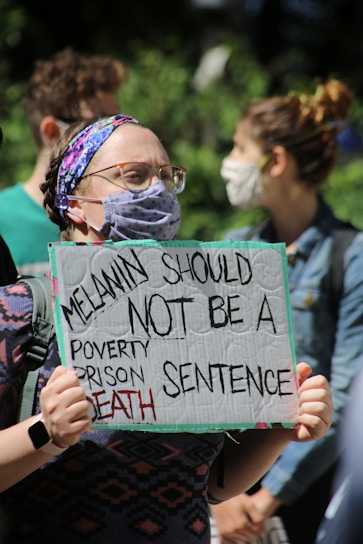 A person wearing a face mask and a colorful headband holds a protest sign that reads 'Melanin should not be a poverty, prison, death sentence'. Others in the background also wear masks, indicating a public gathering or protest.