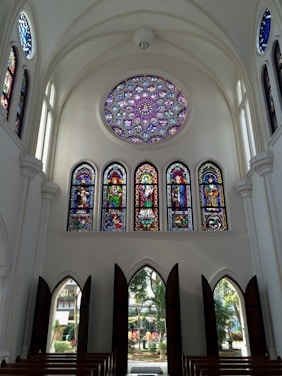 A high-ceilinged church interior with a large, intricate circular stained glass ceiling window and several vertical stained glass windows. The windows depict various religious figures and scenes in vibrant colors. Arched doorways allow natural light to illuminate the space, with wooden benches arranged in rows.