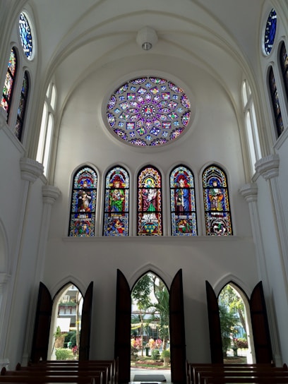 A high-ceilinged church interior with a large, intricate circular stained glass ceiling window and several vertical stained glass windows. The windows depict various religious figures and scenes in vibrant colors. Arched doorways allow natural light to illuminate the space, with wooden benches arranged in rows.