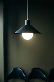 A bright light bulb hanging from the ceiling with a black, conical lampshade in a dimly lit room. The light illuminates a portion of the dark background and part of a row of black chairs.