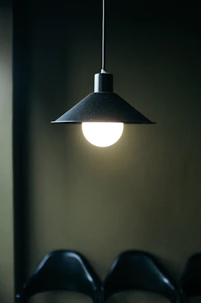 A bright light bulb hanging from the ceiling with a black, conical lampshade in a dimly lit room. The light illuminates a portion of the dark background and part of a row of black chairs.