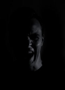 A partially illuminated face of a person in the dark, displaying an expression of anger or frustration with open mouth and furrowed brow. The surrounding darkness emphasizes the intensity of the emotion.
