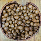 A collection of brown, round seeds or nuts fills a rustic, wooden bowl placed on a wooden surface. The seeds vary slightly in shape and size, and their smooth, glossy surfaces suggest freshness.