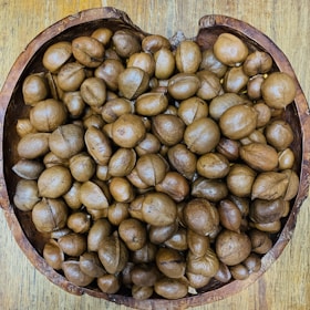 A collection of brown, round seeds or nuts fills a rustic, wooden bowl placed on a wooden surface. The seeds vary slightly in shape and size, and their smooth, glossy surfaces suggest freshness.