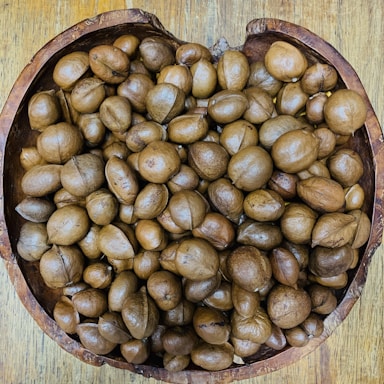 A collection of brown, round seeds or nuts fills a rustic, wooden bowl placed on a wooden surface. The seeds vary slightly in shape and size, and their smooth, glossy surfaces suggest freshness.