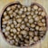 A collection of brown, round seeds or nuts fills a rustic, wooden bowl placed on a wooden surface. The seeds vary slightly in shape and size, and their smooth, glossy surfaces suggest freshness.