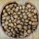 A collection of brown, round seeds or nuts fills a rustic, wooden bowl placed on a wooden surface. The seeds vary slightly in shape and size, and their smooth, glossy surfaces suggest freshness.