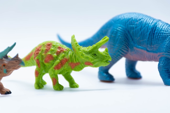 Colorful toy dinosaurs placed in a row against a white background. In the middle, there is a green dinosaur with orange spots and horns. On the right, there is a larger blue dinosaur with a long tail. Partially visible on the left is another dinosaur with a mix of brown and blue colors.