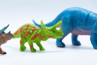 Colorful toy dinosaurs placed in a row against a white background. In the middle, there is a green dinosaur with orange spots and horns. On the right, there is a larger blue dinosaur with a long tail. Partially visible on the left is another dinosaur with a mix of brown and blue colors.