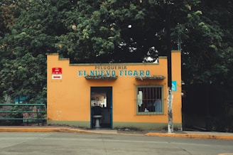 A small barbershop named Peluqueria El Nuevo Figaro with a yellow exterior and a sign with blue lettering. The building is situated along a street with a sidewalk and is surrounded by trees. There is a central entrance with a barber chair visible inside and a window to the right.