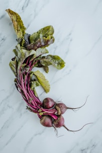 A bundle of fresh beetroots with long, leafy greens is placed on a light marble surface. The roots are deep reddish-purple while the leaves display a mix of green and purple hues, indicating freshness and vibrancy.