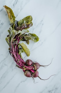 A bundle of fresh beetroots with long, leafy greens is placed on a light marble surface. The roots are deep reddish-purple while the leaves display a mix of green and purple hues, indicating freshness and vibrancy.