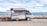 A small mobile store van parked on a concrete surface near a body of water, possibly a sea or ocean. The van has its side panel open, displaying a variety of drinks and snacks arranged in a neat, organized fashion. The background features a clear sky with some clouds and distant mountains across the water. There is also a small red and yellow container next to the van.