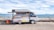 A small mobile store van parked on a concrete surface near a body of water, possibly a sea or ocean. The van has its side panel open, displaying a variety of drinks and snacks arranged in a neat, organized fashion. The background features a clear sky with some clouds and distant mountains across the water. There is also a small red and yellow container next to the van.