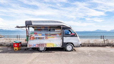 A small mobile store van parked on a concrete surface near a body of water, possibly a sea or ocean. The van has its side panel open, displaying a variety of drinks and snacks arranged in a neat, organized fashion. The background features a clear sky with some clouds and distant mountains across the water. There is also a small red and yellow container next to the van.