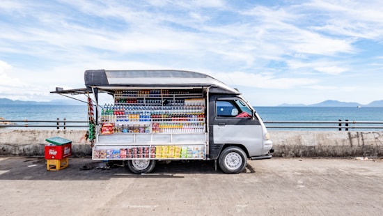 A small mobile store van parked on a concrete surface near a body of water, possibly a sea or ocean. The van has its side panel open, displaying a variety of drinks and snacks arranged in a neat, organized fashion. The background features a clear sky with some clouds and distant mountains across the water. There is also a small red and yellow container next to the van.
