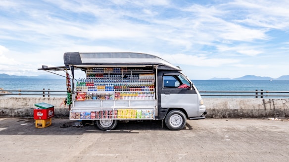 A small mobile store van parked on a concrete surface near a body of water, possibly a sea or ocean. The van has its side panel open, displaying a variety of drinks and snacks arranged in a neat, organized fashion. The background features a clear sky with some clouds and distant mountains across the water. There is also a small red and yellow container next to the van.