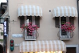 A building facade with two balconies featuring striped awnings and vibrant pink flowers in iron rail planters. There are signs reading 'PIAZZA PORTO VALENTINO' and 'AFFITTASI', indicating a location and something for rent. A traffic light with red and green lights is visible below the balconies. A neon sign, partially visible, hangs to the left.
