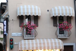 A building facade with two balconies featuring striped awnings and vibrant pink flowers in iron rail planters. There are signs reading 'PIAZZA PORTO VALENTINO' and 'AFFITTASI', indicating a location and something for rent. A traffic light with red and green lights is visible below the balconies. A neon sign, partially visible, hangs to the left.