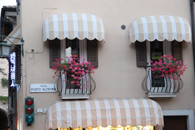 A building facade with two balconies featuring striped awnings and vibrant pink flowers in iron rail planters. There are signs reading 'PIAZZA PORTO VALENTINO' and 'AFFITTASI', indicating a location and something for rent. A traffic light with red and green lights is visible below the balconies. A neon sign, partially visible, hangs to the left.