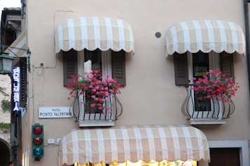 A building facade with two balconies featuring striped awnings and vibrant pink flowers in iron rail planters. There are signs reading 'PIAZZA PORTO VALENTINO' and 'AFFITTASI', indicating a location and something for rent. A traffic light with red and green lights is visible below the balconies. A neon sign, partially visible, hangs to the left.