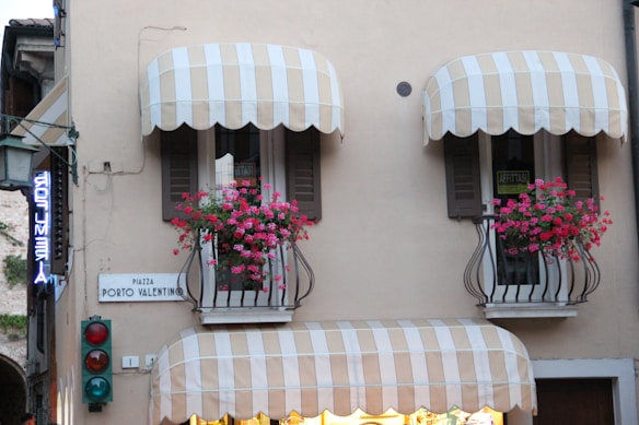 A building facade with two balconies featuring striped awnings and vibrant pink flowers in iron rail planters. There are signs reading 'PIAZZA PORTO VALENTINO' and 'AFFITTASI', indicating a location and something for rent. A traffic light with red and green lights is visible below the balconies. A neon sign, partially visible, hangs to the left.