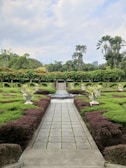 A beautifully landscaped garden features a neatly paved pathway lined with trimmed hedges and symmetrical plant arrangements in large decorative pots. In the background, tall palm trees and vibrant greenery create a lush, tropical atmosphere. The sky is partly cloudy, adding a serene element to the scene.