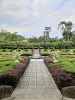 A beautifully landscaped garden features a neatly paved pathway lined with trimmed hedges and symmetrical plant arrangements in large decorative pots. In the background, tall palm trees and vibrant greenery create a lush, tropical atmosphere. The sky is partly cloudy, adding a serene element to the scene.