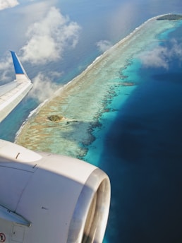 An aerial view of a small island surrounded by turquoise and deep blue waters, seen from an airplane window. The airplane wing and engine are visible, emphasizing travel. The island appears to be part of an atoll or coral reef system, with white sand and green vegetation. There are scattered clouds in the sky above.