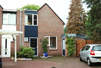 A brick house with a pitched roof and blue accents is situated next to a small driveway. There is a car parked on the driveway, and a garden with various plants, including a tall tree and a bush. A wooden fence encloses the area, adding to the suburban atmosphere.