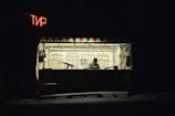 A small shooting range booth with a person sitting inside, surrounded by targets and bottles on shelves. The booth is dimly lit with a neon sign in a foreign script, creating a solitary and somewhat somber atmosphere.