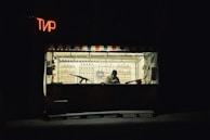 A small shooting range booth with a person sitting inside, surrounded by targets and bottles on shelves. The booth is dimly lit with a neon sign in a foreign script, creating a solitary and somewhat somber atmosphere.