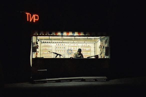 A small shooting range booth with a person sitting inside, surrounded by targets and bottles on shelves. The booth is dimly lit with a neon sign in a foreign script, creating a solitary and somewhat somber atmosphere.