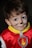 A young child is wearing a red costume with a yellow collar and a badge featuring a flame. The child's face is painted with whiskers and a small black nose, giving them the appearance of a cat or animal character.