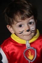 A young child is wearing a red costume with a yellow collar and a badge featuring a flame. The child's face is painted with whiskers and a small black nose, giving them the appearance of a cat or animal character.