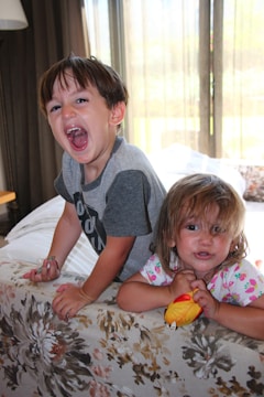 Two children are playfully interacting indoors. The child on the left is laughing energetically, while the child on the right is holding a colorful toy and looking towards the camera. They are leaning on a floral-patterned piece of furniture with sunlight streaming through glass doors in the background.