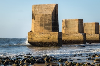 Massive concrete structures are partially submerged in the ocean, with waves gently crashing against them. The tide is low, revealing rocky sections of the shore in the foreground. The image captures a clear blue sky in the background.