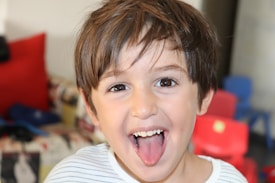A young child with brown hair is smiling widely and playfully sticking out their tongue. The background shows out-of-focus red and blue chairs, suggesting an indoor setting, possibly a play area or classroom.