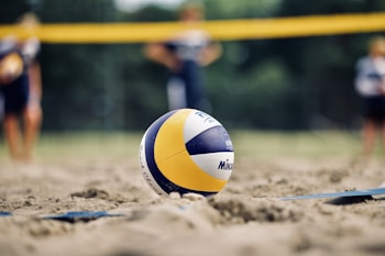 A volleyball rests on a sandy court, with blurred figures in the background suggesting a game setting. The net is visible behind the ball, enhancing the sporty atmosphere.