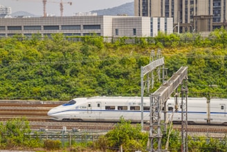 A high-speed train travels on tracks surrounded by lush greenery with buildings in the background. Overhead electrical structures are visible, and there are hills in the distance.