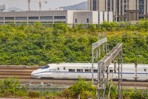 A high-speed train travels on tracks surrounded by lush greenery with buildings in the background. Overhead electrical structures are visible, and there are hills in the distance.