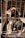 Two puppies are inside a rusty metal cage, with one puppy facing forward and the other looking to the side. They have dark fur with lighter patches and seem to be in a confined space with shadows cast by the cage bars.