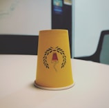 A yellow paper cup is placed upside down on a white table. The cup features a design of a red and black lamp surrounded by a laurel wreath. The background shows a blurry office setting with a chair and some wall postings.