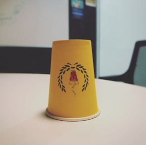 A yellow paper cup is placed upside down on a white table. The cup features a design of a red and black lamp surrounded by a laurel wreath. The background shows a blurry office setting with a chair and some wall postings.