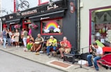 A lively street scene outside a pub, featuring a group of musicians performing with various instruments including guitars and drums. Several people are gathered around, some standing and others sitting, enjoying the live music. The pub has decorative elements like hanging flowers and a colorful rainbow painted on a window.