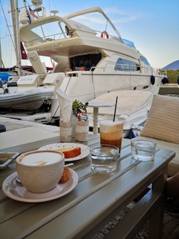A table at a marina is set with a cappuccino, a slice of cake, a glass of cold coffee with a straw, and some water. In the background, a large white yacht is docked alongside other boats. The scene has a leisurely and relaxed atmosphere, with decorative lace and plants adding a touch of elegance.