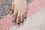 A hand with intricate henna designs covering the fingers and extending to the hand's back. The patterns include floral and abstract motifs, highlighted with intricate detailing. The hand rests on a textured, multi-colored fabric with tones of white, pink, and black.