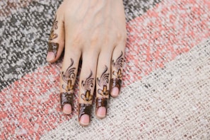 A hand with intricate henna designs covering the fingers and extending to the hand's back. The patterns include floral and abstract motifs, highlighted with intricate detailing. The hand rests on a textured, multi-colored fabric with tones of white, pink, and black.